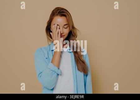 Stanchezza giovane donna che copre un occhio con la mano su sfondo beige che mostra cefalea affaticamento e disagio Foto Stock