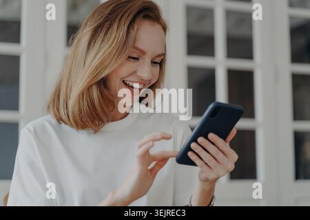 Happy Millennial Woman che usa lo smartphone a casa indossa una camicia bianca e sorride mentre legge il messaggio in Bright Interior Foto Stock