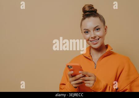 Allegra donna di generazione Z che utilizza lo smartphone sullo sfondo di Beige Studio indossando una felpa arancione e sorridendo durante la messaggistica Foto Stock