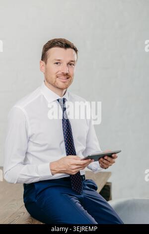 Uomo d'affari con camicia e cravatta bianca tiene il tablet seduto in un moderno interno dell'ufficio Foto Stock
