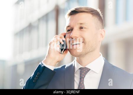 Un uomo d'affari sorridente parla sullo smartphone fuori dal moderno edificio di uffici Foto Stock
