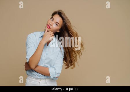 La giovane donna posa con la mano sul mento contro il muro beige dello studio Foto Stock
