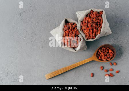 Bacche di goji essiccate in sacchetti di lino e cucchiaio di legno sulla superficie grigia Foto Stock