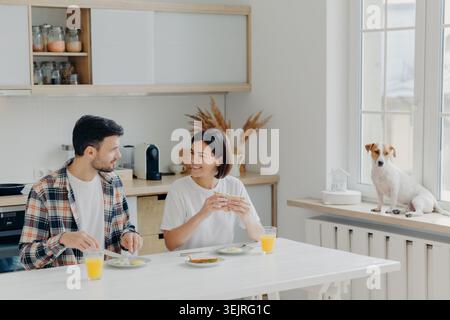 Una giovane coppia fa colazione in una cucina luminosa mentre un piccolo cane si siede accanto alla finestra Foto Stock