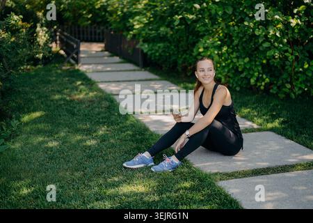 Giovane donna in abbigliamento sportivo seduta sul sentiero del giardino con lo smartphone dopo l'allenamento Foto Stock