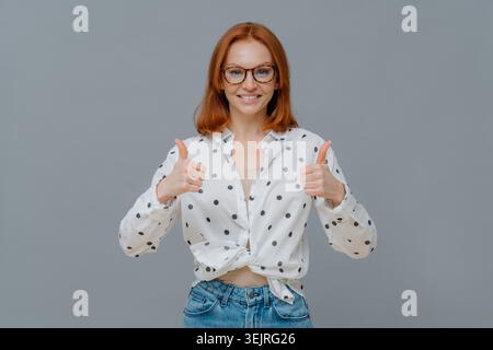 La giovane donna con gli occhiali mostra approvazione con entrambi i pollici in studio Foto Stock