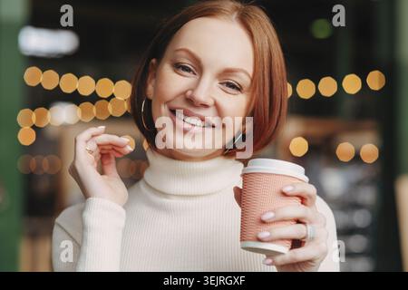 Una donna rossa sorridente tiene una tazza di caffè da asporto in una caffetteria con luci calde Foto Stock