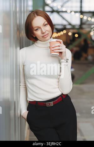 Donna rossa che tiene in mano una tazza di caffè da asporto mentre si appoggia alla finestra del caffè Foto Stock