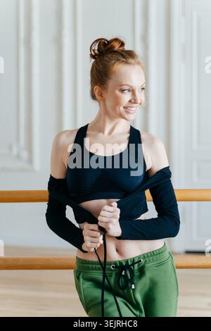 La giovane donna rossa sorridente adatta la giacca sportiva vicino alla sbarra di balletto nello studio di danza Foto Stock