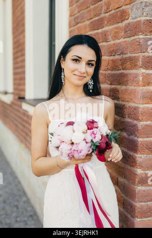 La sposa regala un bouquet di peonie rosa accanto alle pareti di mattoni il giorno del matrimonio Foto Stock