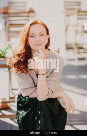 Una donna dai capelli rossi poggia il mento sulla mano mentre si siede nel soleggiato caffè all'aperto Foto Stock