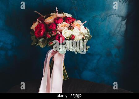 Bouquet nuziale di rose rosse bianche e pesca con foglie di eucalipto e nastri rosa su una parete blu scuro Foto Stock