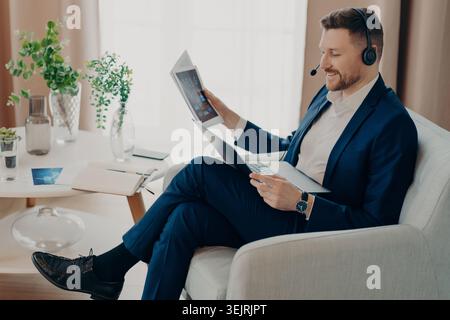 L'uomo d'affari in cuffia esamina i rapporti digitali sul laptop mentre lavora in remoto dal moderno ufficio domestico Foto Stock