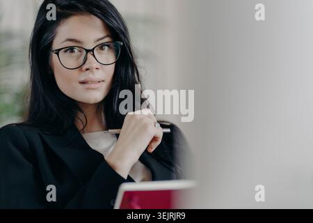 Una giovane donna d'affari che indossa gli occhiali tiene la penna e ascolta durante una riunione professionale Foto Stock