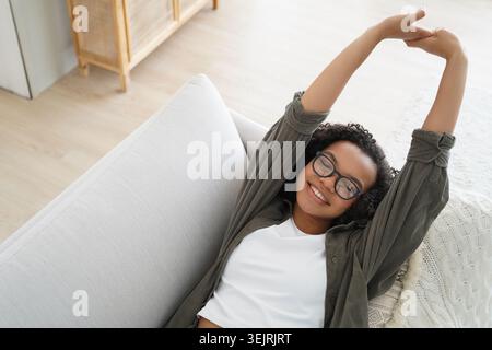 La giovane donna nera che indossa gli occhiali si allunga sul divano in un luminoso salotto di casa Foto Stock