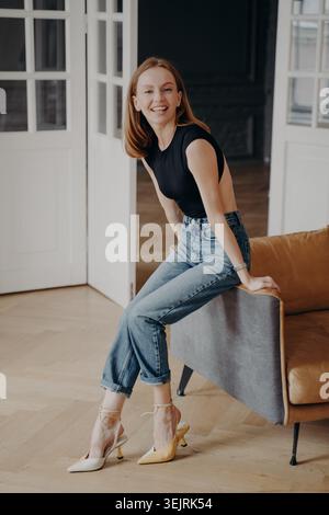 Una giovane donna dai capelli rossi siede sorridente sul divano in eleganti interni moderni Foto Stock