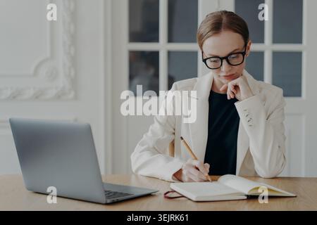 Una donna d'affari scrive appunti sul notebook accanto alla scrivania dell'ufficio Foto Stock