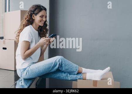Una giovane donna usa lo smartphone mentre si siede su scatole di cartone durante il giorno in movimento Foto Stock