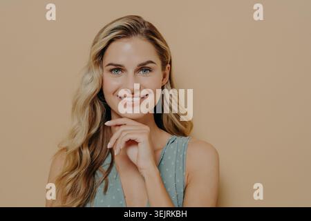 Una donna bionda sorridente tocca il mento mentre posa sullo sfondo beige dello studio Foto Stock