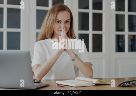 Una donna dai capelli rossi sorride con le mani strette alla scrivania con un laptop e un notebook nell'ufficio domestico Foto Stock