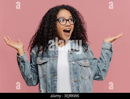 La giovane donna sorpresa con gli occhiali alza le mani mentre si trova contro un muro rosa dello studio Foto Stock