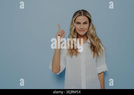La giovane donna bionda punta verso l'alto con l'indice sullo sfondo blu dello studio Foto Stock