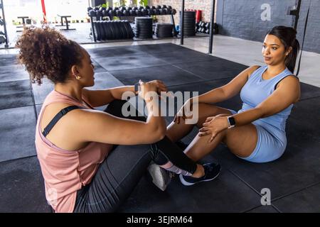 Diverse partner di allenamento femminile che eseguono situp core convenzionati su tappetini da palestra con smartwatch. Duo, abs, crunches, manubri, d Foto Stock