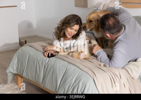 Una coppia sdraiata sul letto accarezzava due Golden retriever su un piumone verde chiaro e uno smartphone nelle vicinanze Foto Stock