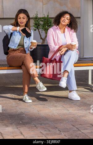 Due donne afroamericane sedute sulla panchina in piazza che controllano lo smartphone e tengono il caffè Foto Stock