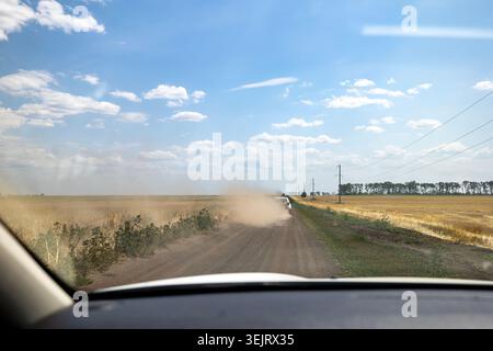 Vista dall'interno di un veicolo che guarda attraverso il parabrezza anteriore un'altra vettura che guida su una strada sterrata polverosa e non asfaltata tra campi agricoli asciutti Foto Stock