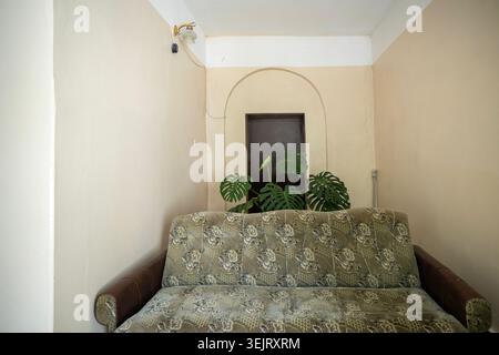 Una vista interna di un divano verde e dorato decorato posizionato di fronte ad una pianta monstera e una porta chiusa marrone scuro posta all'interno di un alcov a parete ad arco Foto Stock