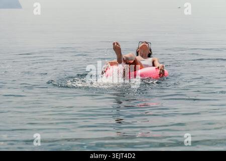 Donna che nuota in mare la giovane signora si rilassa e spruzza acqua mentre galleggia su un anello gonfiabile rosa nell'oceano calmo durante le vacanze estive Foto Stock
