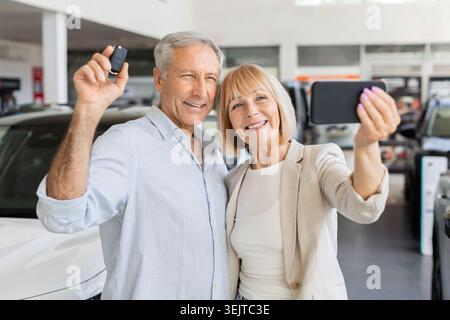 Coppia anziana che scatta selfie con lo smartphone e la chiave dell'auto presso lo showroom della concessionaria Foto Stock
