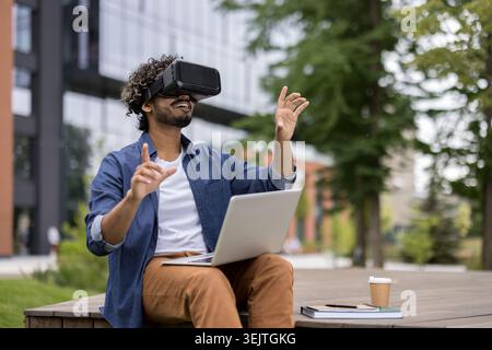 Giovane uomo che indossa un visore vr e tiene in mano un laptop, seduto all'aperto e interagisce con un ambiente virtuale utilizzando i gesti delle mani, sperimentando la realtà aumentata e l'innovazione digitale Foto Stock
