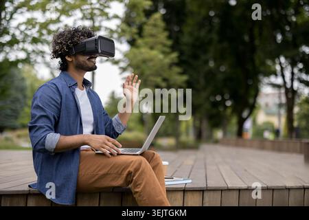 Giovane uomo che indossa un visore per la realtà virtuale e utilizza un notebook seduto all'aperto su un ponte di legno in un parco, coinvolgendo i contenuti di realtà virtuale e le tecnologie emergenti Foto Stock