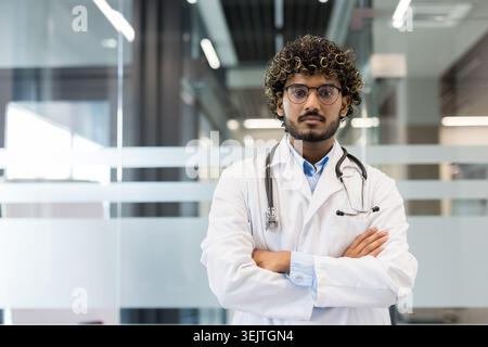 Giovane indiano maschio medico in bianco cappotto con stetoscopio, braccia incrociate, sicuro di espressione seria, in piedi in moderno ospedale interno, ritratto professionale, vista frontale Foto Stock