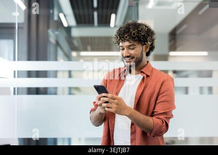 Giovane e affascinante uomo con capelli ricci sorridenti e messaggi sullo smartphone, che gode di comunicazioni mobili e tecnologia moderna in un ambiente d'ufficio moderno e luminoso Foto Stock