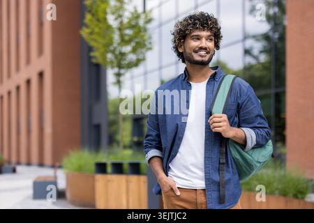 Sorridente giovane studente indiano con capelli ricci che cammina in sicurezza in un moderno campus universitario, portando con sé uno zaino e guardando lontano, rappresentando l'istruzione superiore, il successo e un futuro luminoso Foto Stock