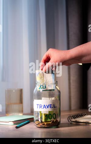 Versare il denaro contante in un vaso di vetro sul tavolo. Primo piano delle dita che inseriscono il denaro in un contenitore trasparente per viaggiare. Pianificazione finanziaria e. Foto Stock