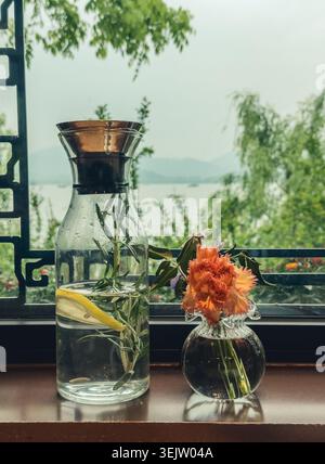 Caraffa limpida con acqua di limone rosmarino e fiori d'arancio che decorano un davanzale in legno che si affaccia su un tranquillo lago Foto Stock