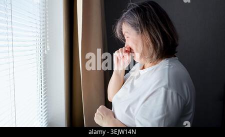 Donna anziana che piange con la mano sul viso, si sente isolata, triste e soffre di depressione mentre sta sola vicino a una finestra in una luce soffusa Foto Stock