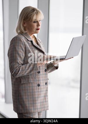 Donna d'affari matura in una tuta a plaid che si concentra profondamente su un computer portatile, gestendo diligentemente le attività professionali e connettendosi con la tecnologia tramite un Foto Stock