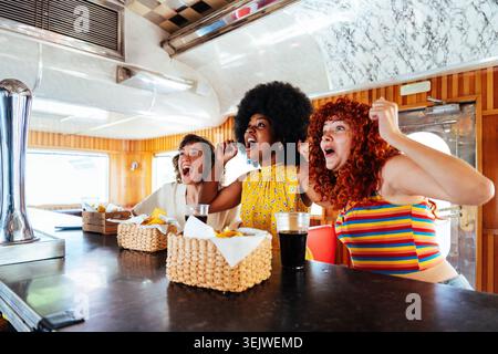 Gruppo multietnico di giovani amici felici che si uniscono al ristorante vintage Burger House - tre eccitanti donne multietniche che esultano e legano al A. Foto Stock