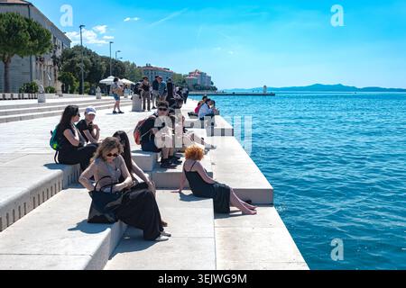 I turisti si rilassano sui gradini di pietra bianca del Sea Organ a Zara, Croazia, affacciati sul vivace mare Adriatico blu in una giornata di sole. Foto Stock