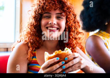 Gruppo multietnico di giovani amici felici che si uniscono al ristorante vintage Burger House - donna felice con capelli ricci rossi che mangiano un hamburger e laughi Foto Stock
