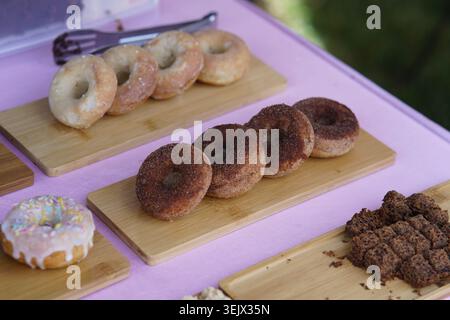 Ciambelle fresche con zucchero alla cannella e colorate spruzzette esposte su tavole di legno al soleggiato chiosco estivo dei cibi del festival. Tonbridge Foto Stock