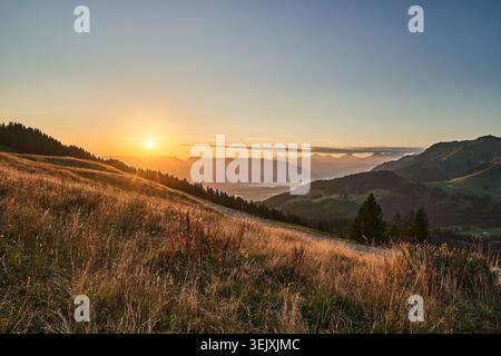 Il tramonto bagna un prato alpino e montagne ondulate con una calda luce dorata e una strada tortuosa. Foto Stock