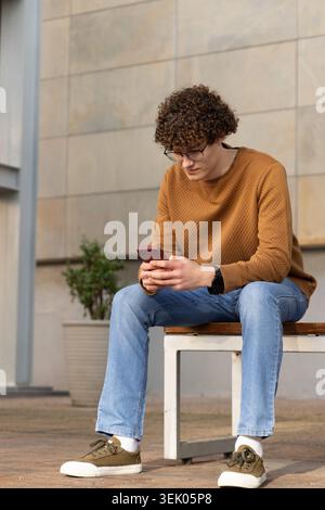 Uomo di vent'anni seduto su una panchina di legno che regge uno smartphone rosso nel cortile urbano, indossando occhiali Foto Stock