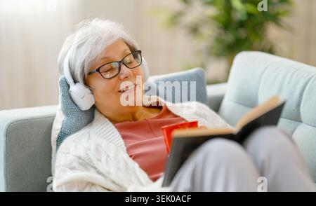 Donna anziana allegra con capelli e occhiali grigi, comodamente sdraiata su un morbido divano e con una comoda maglieria. È immersa nel relax, listeni Foto Stock