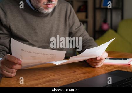 Uomo maturo che tiene due documenti e li confronta a casa. Pianificazione del bilancio e concetto di decisione finanziaria. Luce calda, composizione in primo piano, per interni Foto Stock
